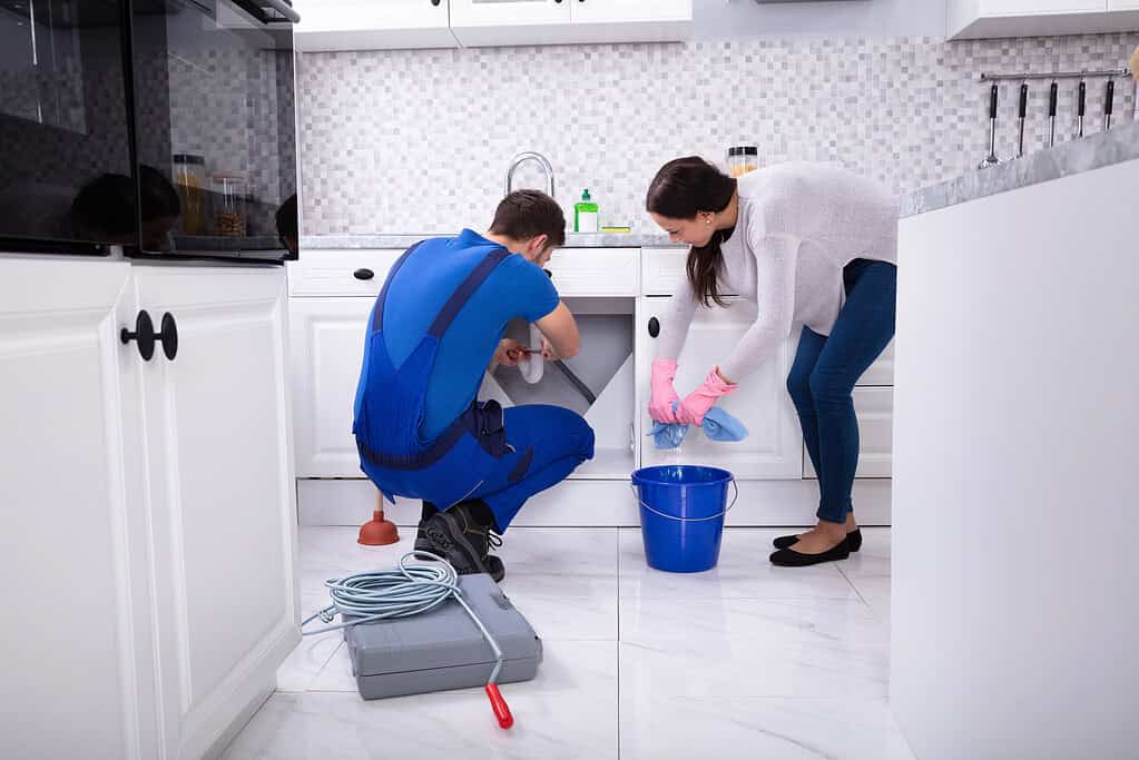 A professional plumber in a clean, modern workshop, wearing a blue uniform and safety goggles, examines a complex piping system with a specialized drain cleaning tool in hand. In the foreground, a shiny metal drain auger lies on a tool bench, indicating readiness for action. The middle-ground features various plumbing tools and equipment neatly arranged on shelves, with some pipes and fittings visible. In the background, a large window casts soft daylight into the space, creating an atmosphere of cleanliness and efficiency. The overall mood is one of expertise and professionalism, highlighting the thorough and dedicated approach to tackling stubborn clogs. The scene captures a moment before the plumber begins the cleaning process, emphasizing the need for professional intervention in serious cases of pipe blockage. Professionelle Rohrreinigung